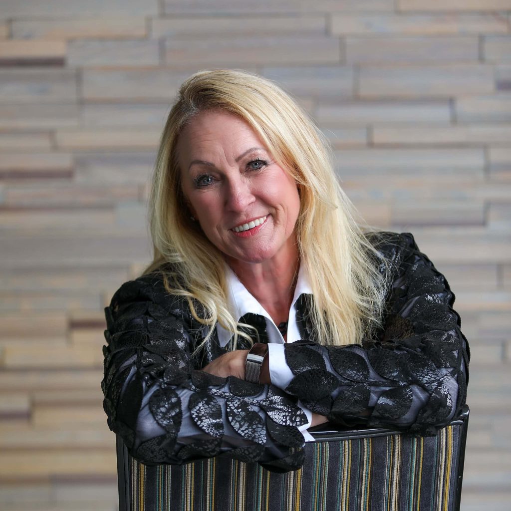 Professional woman smiling, seated in modern office with wood accent wall, showcasing confident leadership and expertise in technology and business solutions.